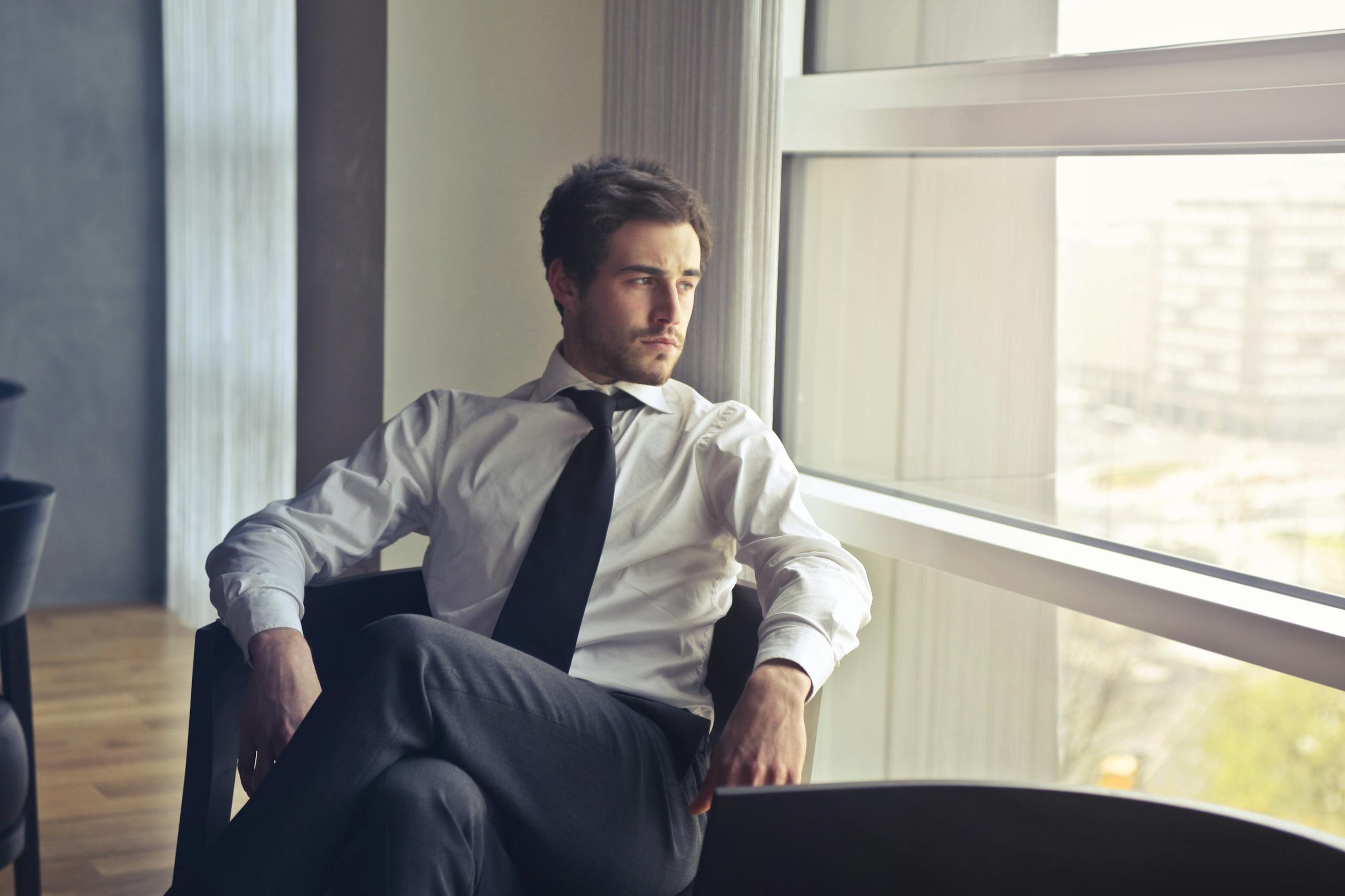 Young businessman in formal attire sitting and looking out of modern office window, contemplating.